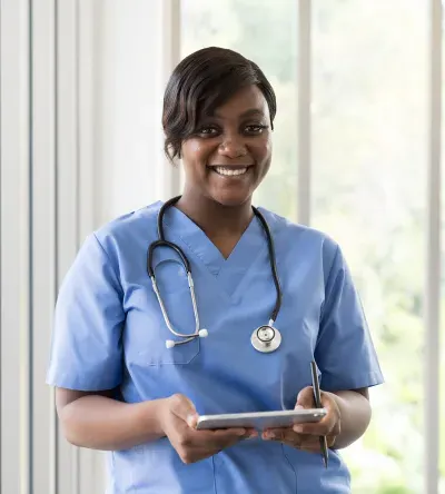 Smiling medical assistant in blue scrubs with a stethoscope, holding a tablet indoors.