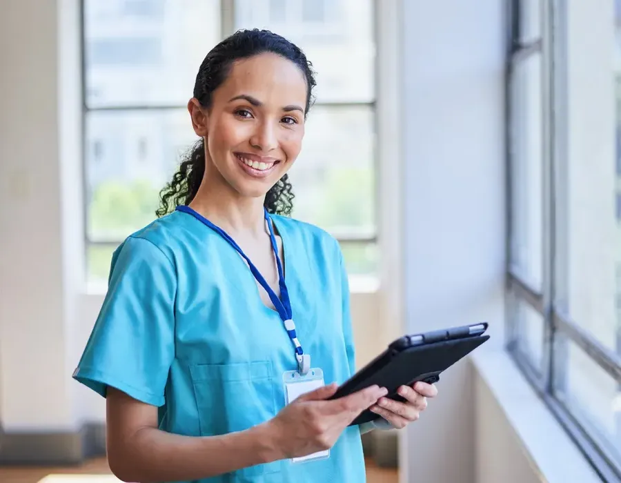 Practical nurse with prior medical assistant experience wearing blue scrubs holding clipboard in medical clinic